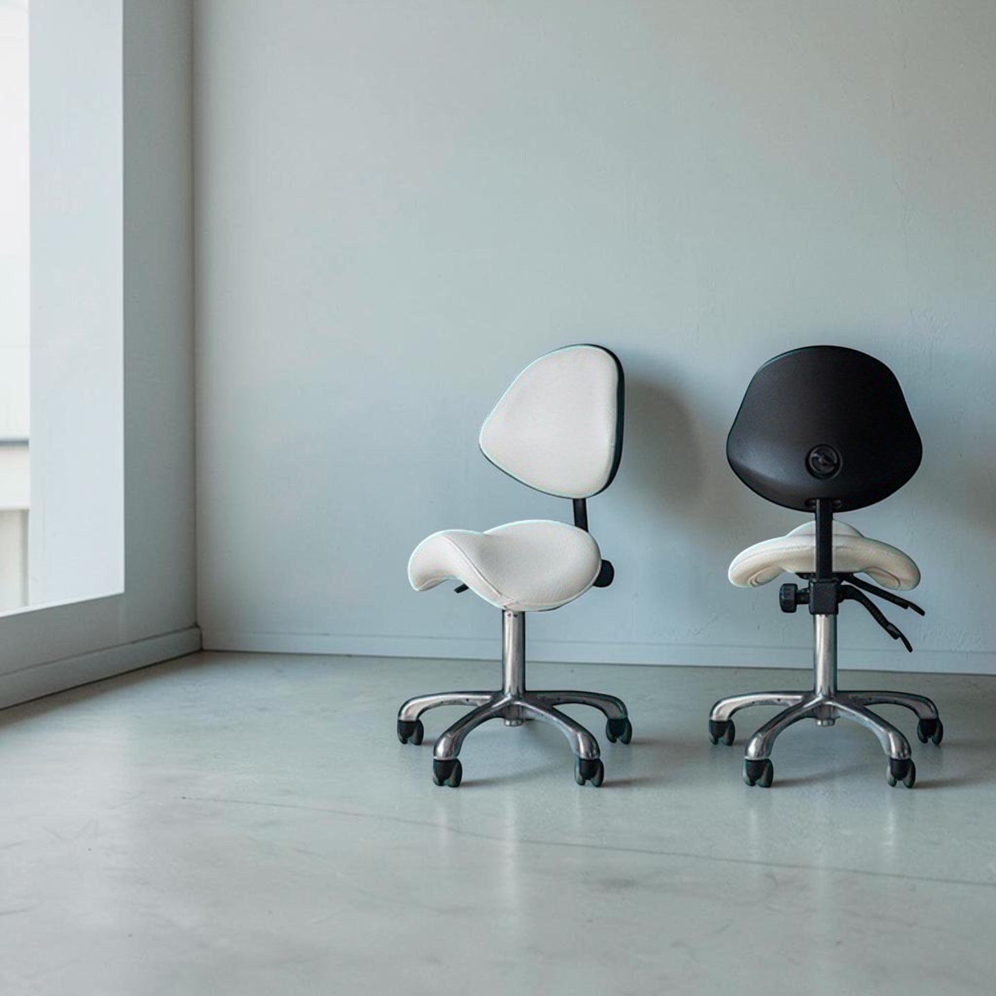 MK BEAUTY BED ergonomic saddle stools with backrests in white and black, featuring chrome bases and rolling casters.