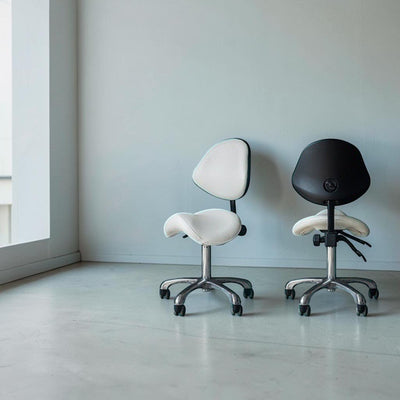 MK BEAUTY BED ergonomic saddle stools with backrests in white and black, featuring chrome bases and rolling casters.